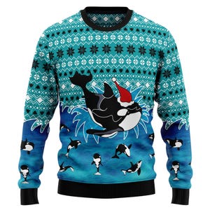 May include: A teal and blue Christmas sweater featuring a large orca wearing a Santa hat, surrounded by smaller orcas and waves. The sweater has a patterned design with snowflakes and geometric shapes, and black cuffs and hem.