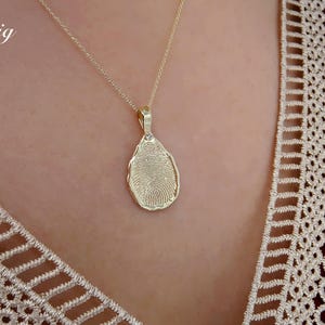 May include: A gold-colored pendant necklace featuring a fingerprint design. The teardrop-shaped pendant hangs from a delicate chain. The word "Big" is written in white script in the upper left corner. The necklace is worn over a cream-colored lace top.