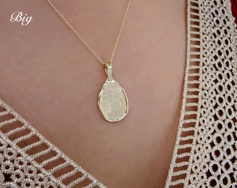 May include: A gold-colored pendant necklace featuring a fingerprint design. The teardrop-shaped pendant hangs from a delicate chain. The word "Big" is written in white script in the upper left corner. The necklace is worn over a cream-colored lace top.