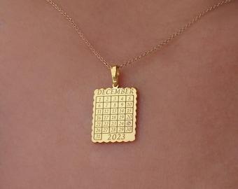 May include: A gold-colored pendant necklace featuring a calendar design for December 2023. The pendant is square with a scalloped edge and displays the month and year, along with the days of the month. The necklace chain is also gold.