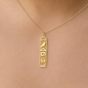 Puede incluir: Un collar de tono dorado con un colgante vertical de forma ovalada alargada. El colgante está diseñado como un cartucho que contiene una serie de símbolos jeroglíficos egipcios recortados, incluyendo un pájaro, un semicírculo y formas geométricas.