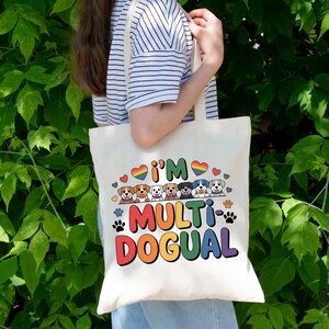 May include: Cream-colored tote bag with the text "I'M MULTI-DOGUAL" in rainbow colors, featuring cartoon dog illustrations and paw prints. The bag has a simple design and is held against a backdrop of green foliage.