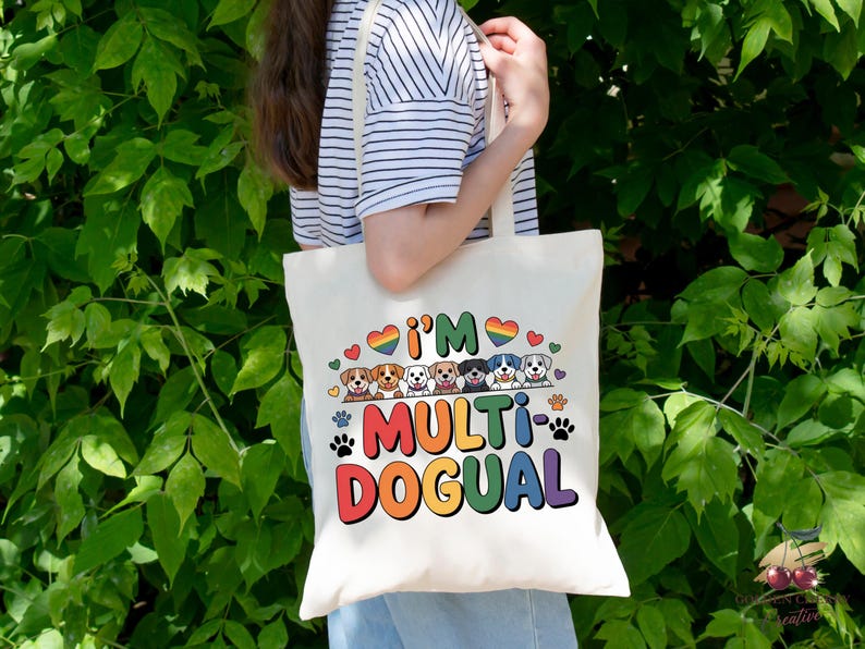 May include: Cream-colored tote bag with the text "I'M MULTI-DOGUAL" in rainbow colors, featuring cartoon dog illustrations and paw prints. The bag has a simple design and is held against a backdrop of green foliage.