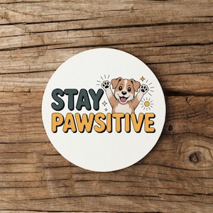 May include: A round, white coaster with the words "STAY PAWSITIVE" in teal and orange lettering. A cartoon dog with its paws up is in the center. The coaster is on a wooden surface.