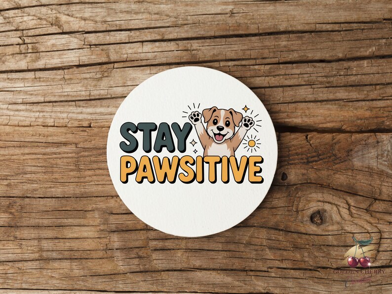 May include: A round, white coaster with the words "STAY PAWSITIVE" in teal and orange lettering. A cartoon dog with its paws up is in the center. The coaster is on a wooden surface.