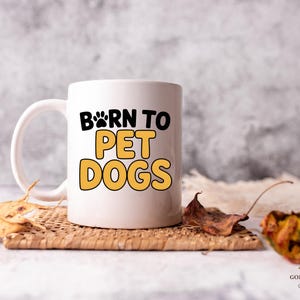 May include: White ceramic mug with the text "BORN TO PET DOGS" in black and yellow. The mug has a paw print design and is sitting on a woven coaster with dried leaves.