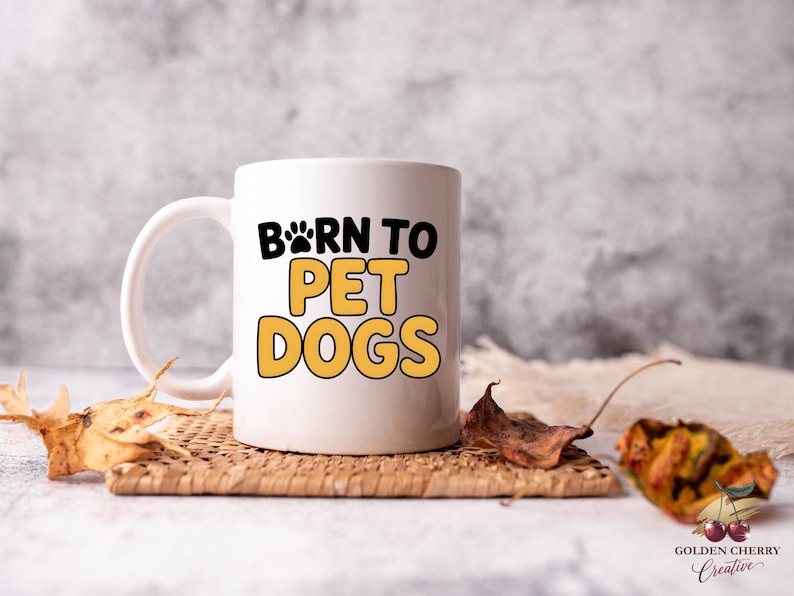 May include: White ceramic mug with the text "BORN TO PET DOGS" in black and yellow. The mug has a paw print design and is sitting on a woven coaster with dried leaves.