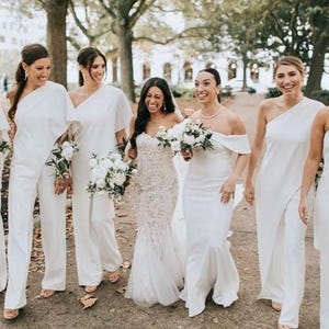 May include: A group of people, including a bride, are walking outdoors. The bride wears a white lace gown, while the others wear white outfits, including jumpsuits and dresses. Each person carries a bouquet of white flowers. The setting is a tree-lined path.