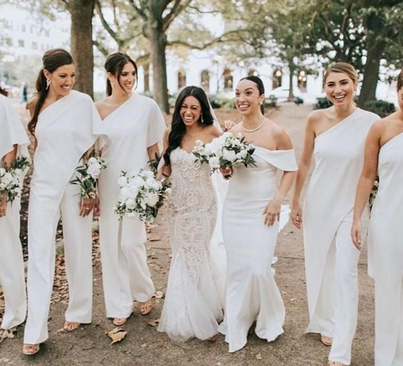 May include: A group of people, including a bride, are walking outdoors. The bride wears a white lace gown, while the others wear white outfits, including jumpsuits and dresses. Each person carries a bouquet of white flowers. The setting is a tree-lined path.