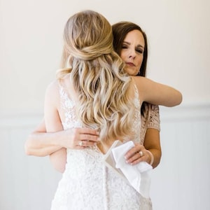 May include: A bride in a white lace wedding dress is embraced by another person. The bride's blonde hair is styled in loose waves. The other person is wearing a pink lace top and holding a white handkerchief.