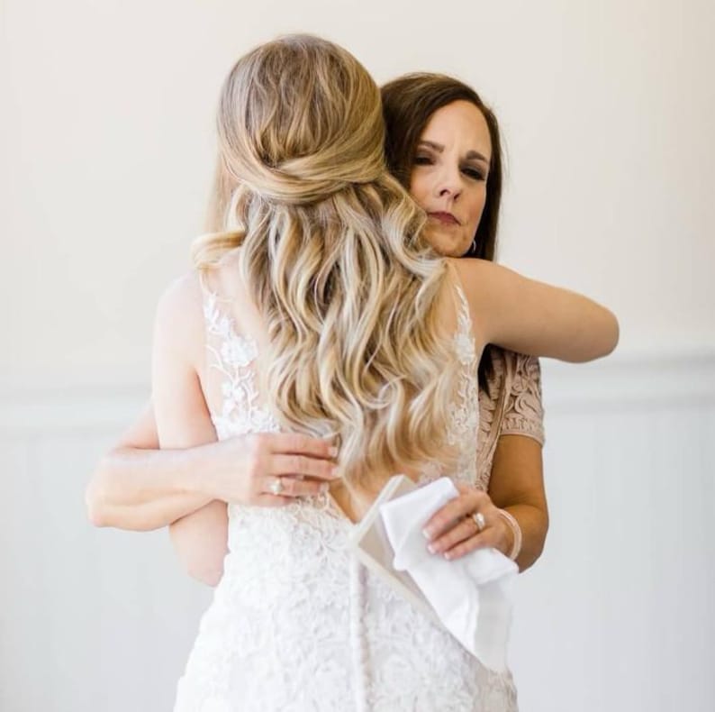 May include: A bride in a white lace wedding dress is embraced by another person. The bride's blonde hair is styled in loose waves. The other person is wearing a pink lace top and holding a white handkerchief.