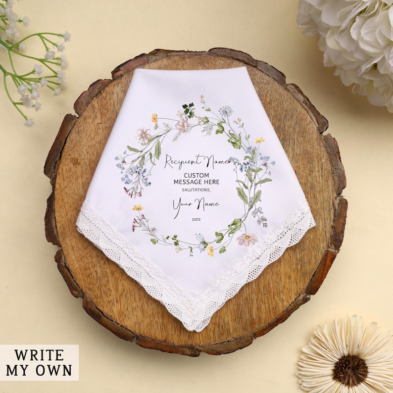 May include: White handkerchief with a floral wreath design and lace trim. The handkerchief has text that reads "Recipient Name", "Custom Message Here", "Your Name", and "Date". The handkerchief is on a wooden surface.