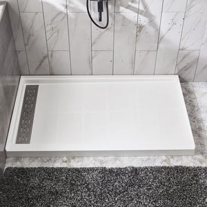 May include: A white rectangular shower base with a stainless steel drain cover. The shower base is set against a marble-tiled wall and a gray, shaggy bath mat. A shower head is visible above.