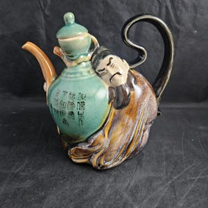 May include: A ceramic teapot with a green glaze and a brown, sculpted figure of a man with a beard. The teapot has a black handle and a small lid. The teapot has Chinese characters on the side.