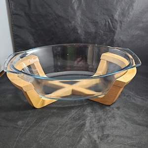 May include: A clear glass baking dish with a light blue rim, set in a light brown wooden cradle. The dish has a curved oval shape and handles on either side. The wooden cradle has an X-shaped design.