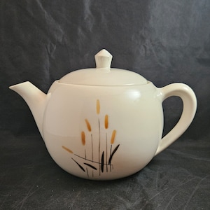May include: White ceramic teapot with a lid and a handle. The teapot has a design of brown and gold stylized grass on the side.