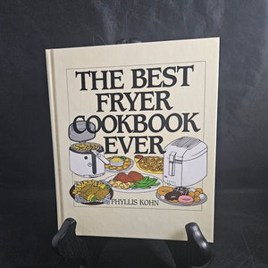 May include: A beige cookbook with the title "The Best Fryer Cookbook Ever" in black text. The cover features illustrations of a deep fryer, french fries, onion rings, donuts, and other fried foods. The author's name, "Phyllis Kohn", is printed in black text at the bottom of the cover.