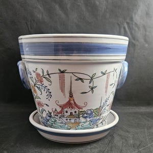 May include: A white ceramic planter with blue and gold trim. The planter features a hand-painted design of a pagoda with flowers and vines. The planter has two blue handles and a matching saucer.
