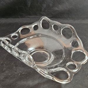 May include: A clear glass bowl with a scalloped edge and a decorative design of small circles around the rim. The bowl is designed to hold small items like candy or jewelry.