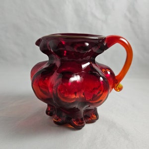 May include: A small, red glass pitcher with a bulbous body and a flared rim. The pitcher has a single, orange handle and a textured surface.