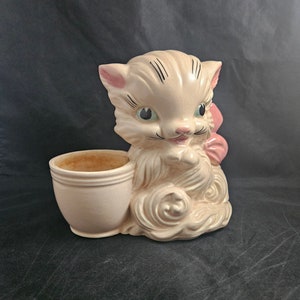 May include: A ceramic planter shaped like a white kitten with a pink bow. The kitten has black eyes and a pink nose. The planter has a separate pot for planting.