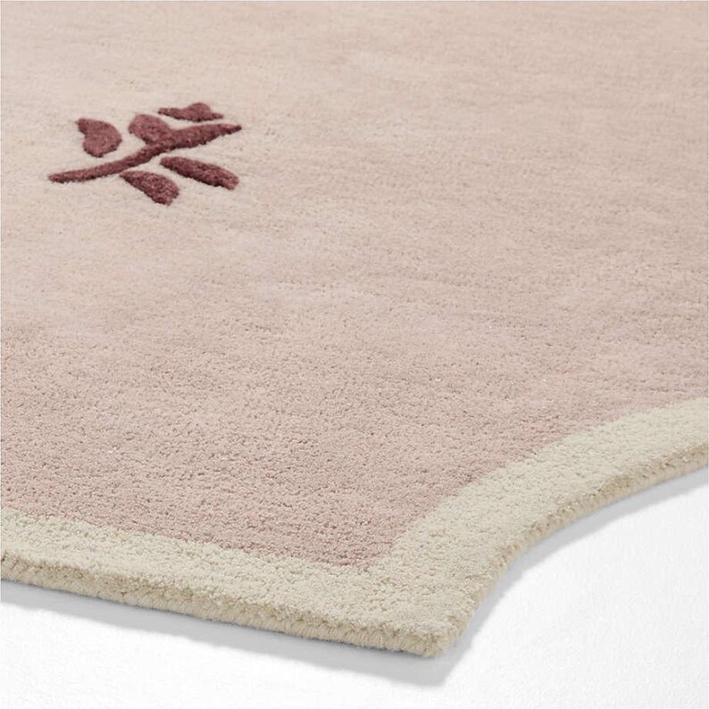 Puede incluir: Primer plano de una alfombra rosa claro y suave con un dise&ntilde;o floral rosa oscuro en la esquina superior izquierda. La alfombra tiene un borde color crema. La alfombra est&aacute; hecha de un material afelpado.