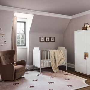 Puede incluir: Un cuarto de beb&eacute; con una cuna blanca, un sill&oacute;n marr&oacute;n y un armario blanco. La habitaci&oacute;n tiene un esquema de color lila claro con una alfombra estampada y tres cuadros enmarcados en la pared. Un m&oacute;vil cuelga del techo.