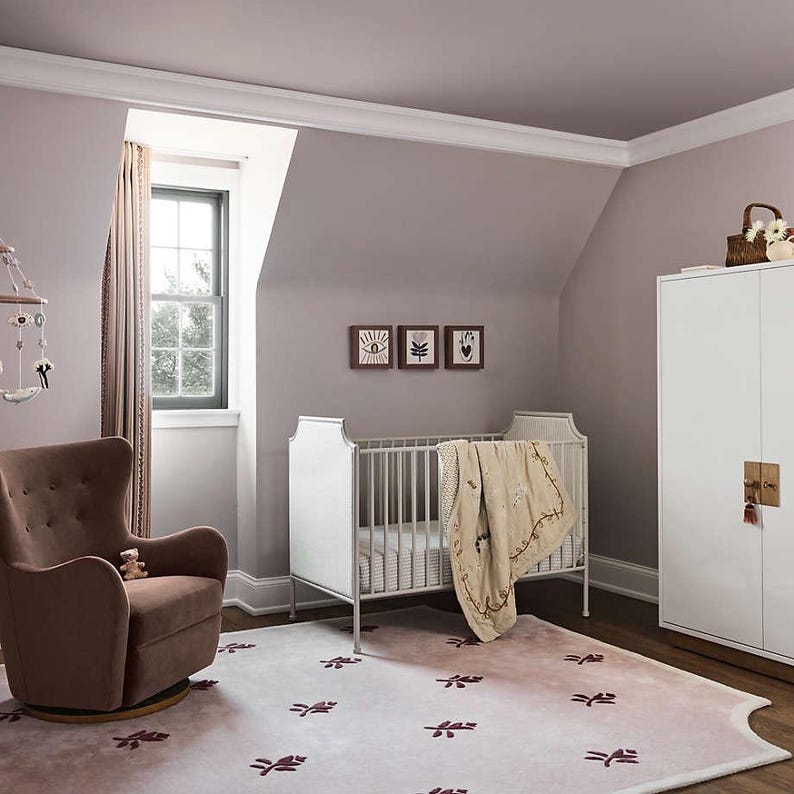 Puede incluir: Un cuarto de beb&eacute; con una cuna blanca, un sill&oacute;n marr&oacute;n y un armario blanco. La habitaci&oacute;n tiene un esquema de color lila claro con una alfombra estampada y tres cuadros enmarcados en la pared. Un m&oacute;vil cuelga del techo.