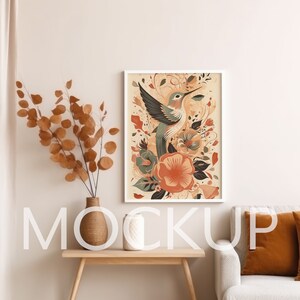 Puede incluir: Una impresión enmarcada de un colibrí con flores y hojas coloridas. La impresión está en un marco blanco y está colgada en una pared blanca. Hay un jarrón con ramas secas sobre una mesa de madera delante de la impresión.