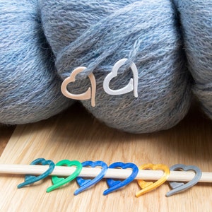 Op de afbeelding: Drie bollen blauwe wol met witte hartvormige steekmarkeerders. Een houten breinaald met kleurrijke hartvormige steekmarkeerders.