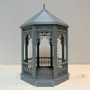 Puede incluir: Un cenador gris en miniatura con intrincados detalles calados. El cenador presenta un techo hexagonal y una barandilla decorativa en la base. El suelo interior tiene un patrón de ladrillos. La estructura es de un material gris sólido.