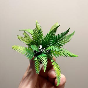 May include: A small, artificial fern plant with vibrant green leaves. The fern has a detailed, layered design, with each frond showing intricate patterns. The plant is potted in a small, dark container, creating a natural aesthetic. The fern is held in a hand.