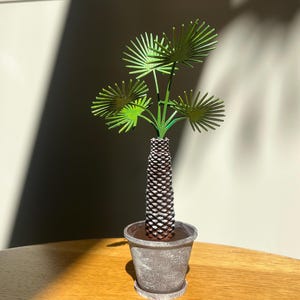 Puede incluir: Una escultura decorativa de palmera en una maceta gris. La palmera tiene hojas verdes y un tronco marrón con un patrón texturizado. La maceta es redonda con un platillo a juego. La escultura está sobre una mesa de madera.