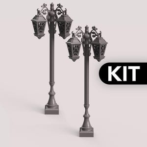 Può includere: Due lampioni grigi e ornati, ciascuno con tre lanterne. Le lampade presentano decorazioni a volute e sono montate su basi quadrate. La parola "KIT" è visualizzata in un riquadro nero.