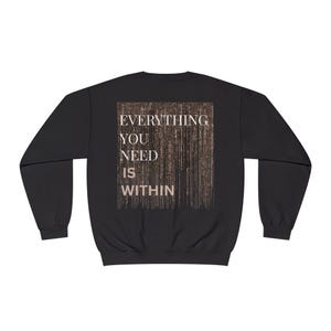 Könnte beinhalten: Schwarzes Sweatshirt mit dem Text "EVERYTHING YOU NEED IS WITHIN" in einem rechteckigen Design auf dem Rücken. Der Text ist in einer Serifenschrift, und der Hintergrund hat ein braunes, strukturiertes Aussehen.