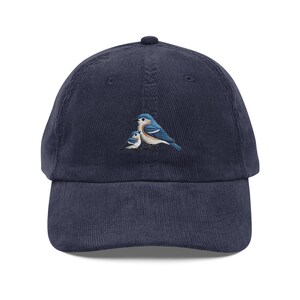 Peut inclure: Une casquette de baseball en velours côtelé bleu marine avec un motif brodé de deux oiseaux bleus. Les oiseaux sont perchés ensemble, le plus grand à droite et le plus petit à gauche.