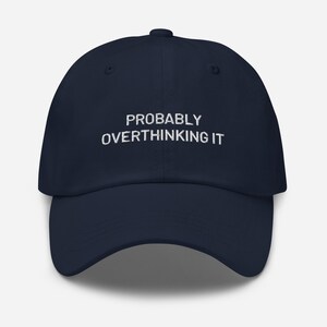 Puede incluir: Gorra de béisbol azul marino con el texto bordado "PROBABLY OVERTHINKING IT" en blanco. La gorra tiene una visera curva y un cierre de correa de tela en la parte trasera. Un accesorio casual.