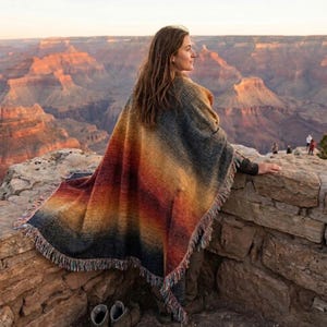 May include: A colorful poncho with a fringed edge, featuring a gradient of colors from dark blue to orange and yellow. The poncho is draped over a person, with a scenic canyon landscape in the background.