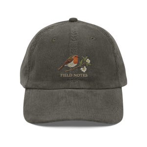 Puede incluir: Gorra de béisbol de pana verde oliva con un diseño bordado de un petirrojo posado en una rama florida. El texto "FIELD NOTES" está bordado debajo. La gorra tiene una visera curva y una correa ajustable.