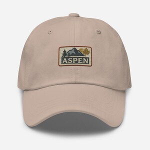 Könnte beinhalten: Eine beige Baseballkappe mit einem gestickten Aufnäher, der eine Bergszene und das Wort "ASPEN" in einem Vintage-Stil zeigt.