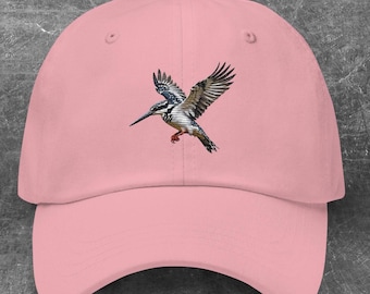 May include: A light pink baseball cap featuring an embroidered image of a bird in flight. The bird has black, white, and brown feathers, with a long beak and red feet. The cap has a curved brim and a button on top.