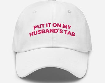 May include: White baseball cap with the phrase "PUT IT ON MY HUSBAND'S TAB" embroidered in pink. The cap has a curved brim and a classic six-panel design. A casual accessory for everyday wear.