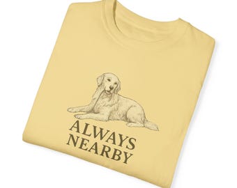 Puede incluir: Una camiseta amarilla con un dibujo de un golden retriever tumbado y el texto "ALWAYS NEARBY" impreso en la parte delantera.