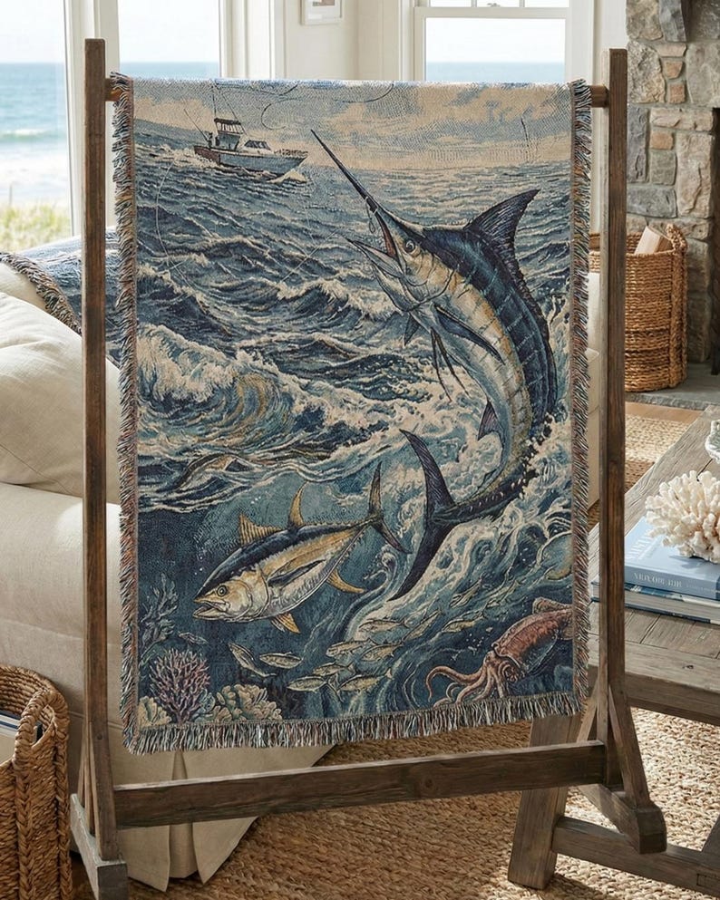 May include: A woven tapestry featuring a marine scene with a marlin leaping from the ocean, a tuna, and a boat in the distance. The tapestry is displayed on a wooden stand, showcasing a nautical theme with shades of blue, beige, and brown.