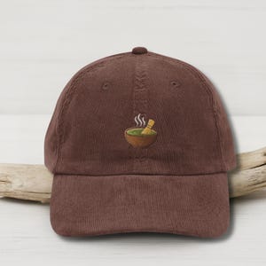 May include: Brown corduroy baseball cap featuring an embroidered matcha tea bowl and whisk design. The bowl is green with steam rising, and the whisk is tan. The cap has a curved peak and a classic six-panel design.