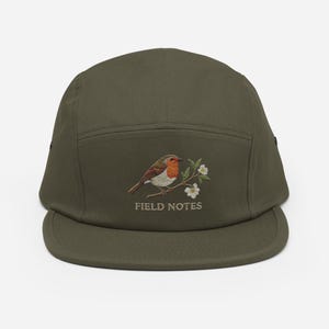 May include: Olive green five-panel cap with a detailed embroidered design. The design features a robin perched on a branch with white flowers, and the text "FIELD NOTES" below. The cap has a flat brim.