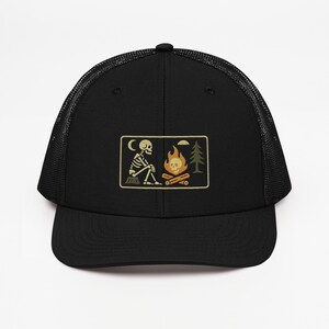 May include: Black trucker hat with a mesh back and a curved brim. The front features an embroidered patch with a skeleton sitting by a campfire under a crescent moon and a pine tree. The patch has a tan border.