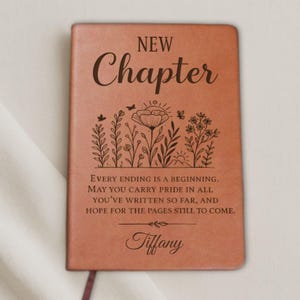 May include: A brown leather journal with the words "NEW Chapter" engraved on the cover. Below the title is a floral design and a quote. The name "Tiffany" is also engraved on the cover. A maroon ribbon bookmark is visible.