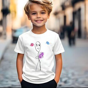 May include: White t-shirt featuring a whimsical stick figure design. The illustration includes a smiling face, a purple ball, and red and blue scribbles. The shirt is a classic crew neck style.