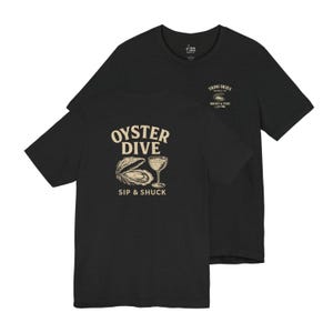Op de afbeelding: Zwart T-shirt met een afbeelding van een oester en een cocktailglas met de tekst "Oyster Dive Sip & Shuck". De zak op het shirt heeft een klein logo met de tekst "Trini Skies Supply Co. Briny & Fine".